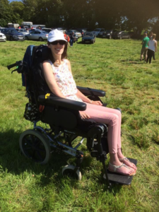 The image depicts a woman sitting in a wheelchair on a grassy field outdoors. She is wearing sunglasses and is dressed in casual clothing. Surrounding her are elements of nature, including trees and open land. The wheelchair features tires typical of a vehicle designed for mobility.