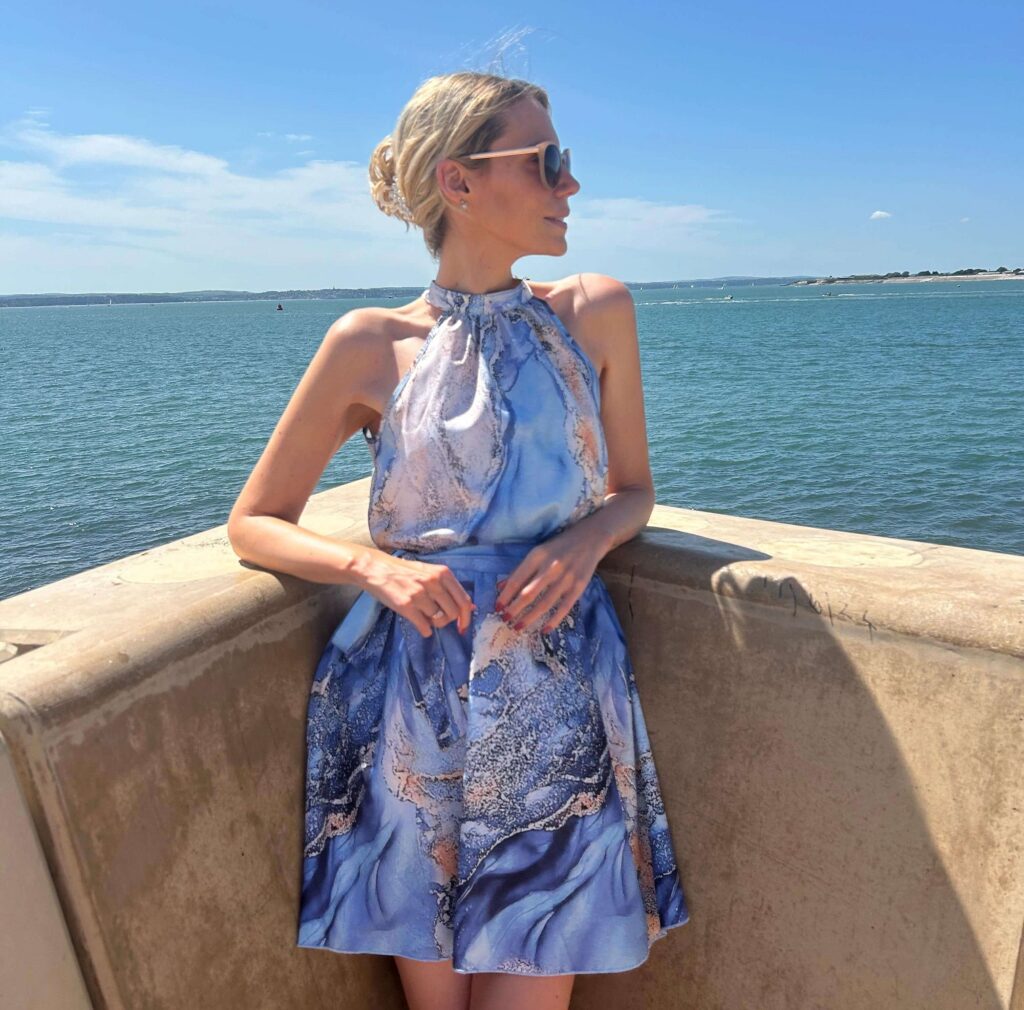 The image depicts a woman wearing a blue dress, enjoying a sunny day outdoors at the beach. She is accessorized with sunglasses and is likely on vacation, with the sky and water in the background. The setting suggests a summery atmosphere, highlighting her stylish fashion choice.