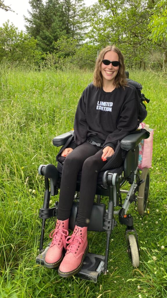 A young woman in a wheelchair wearing pink glitter boots and sunglasses smiles at the camera while sitting in a grassy field. She is wearing a black sweatshirt that says "limited edition" on it. The photo is taken from a low angle, giving the viewer the feeling that they are looking up at the woman.