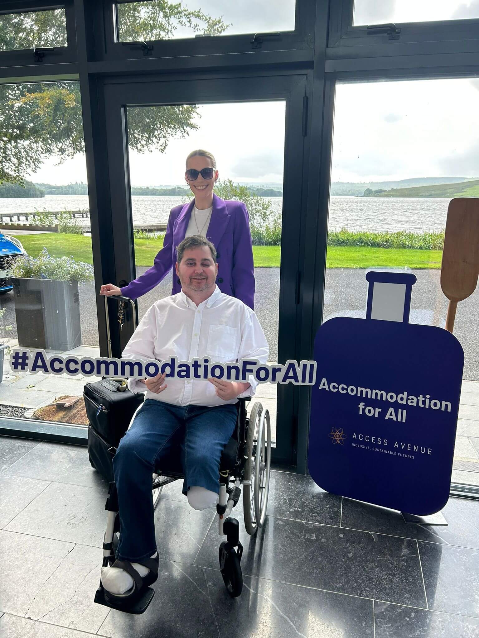 "Inside a building with large windows overlooking a lake. A woman in a purple blazer stands behind a man in a wheelchair. The man wears a white shirt. In front of them is a sign reading '#AccommodationForAll'. To the right is a blue board with 'Accommodation for All' and 'Access Avenue' written on it. The background shows a lake view with grassy areas."