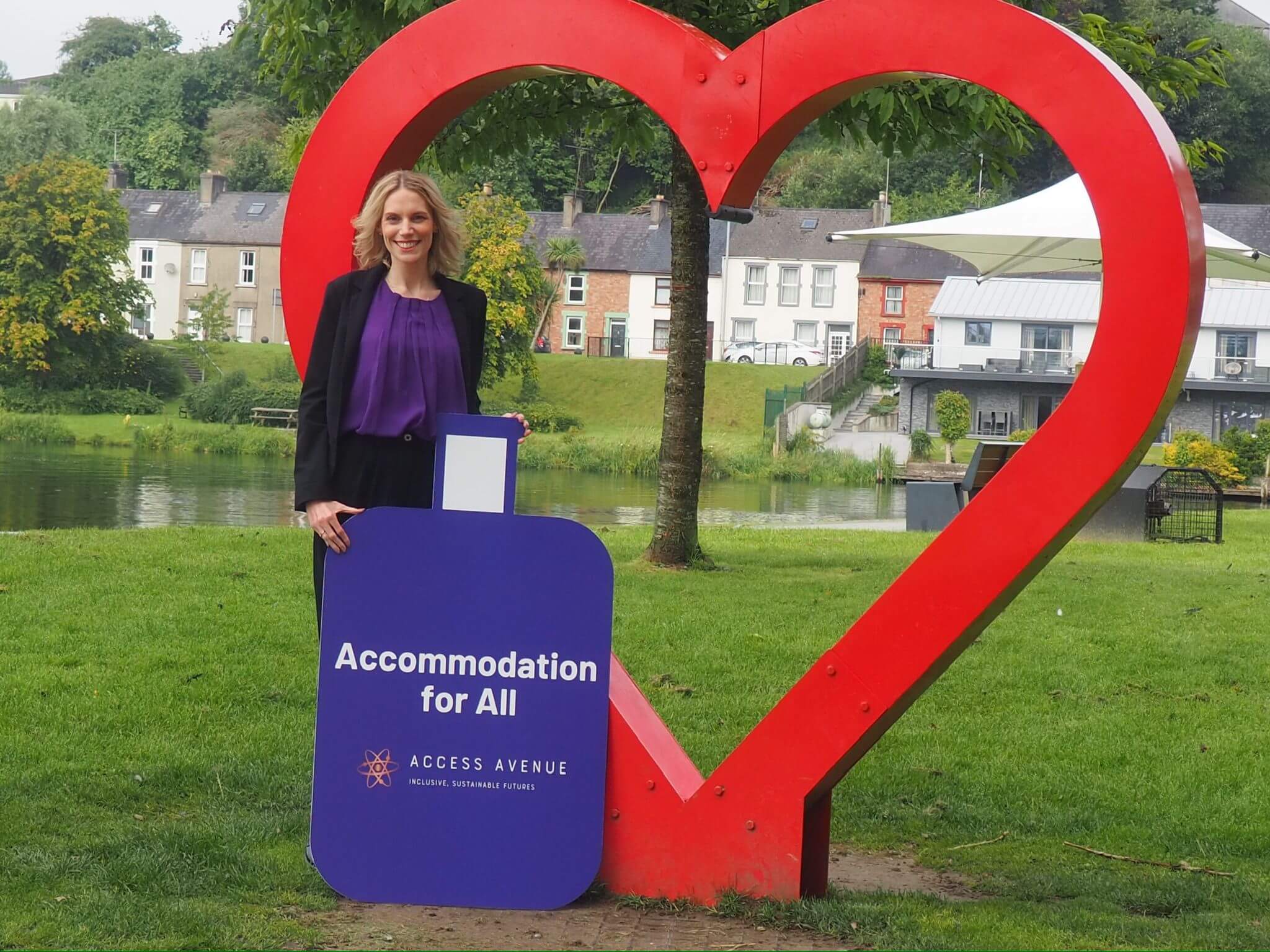 "A smiling blonde woman in a purple top and black blazer stands in a grassy area holding a purple sign that reads 'Accommodation for All' with the Access Avenue logo. She's positioned next to a large red heart-shaped sculpture. In the background, there's a river or canal and a row of colorful houses. The scene suggests a picturesque small town or village setting."