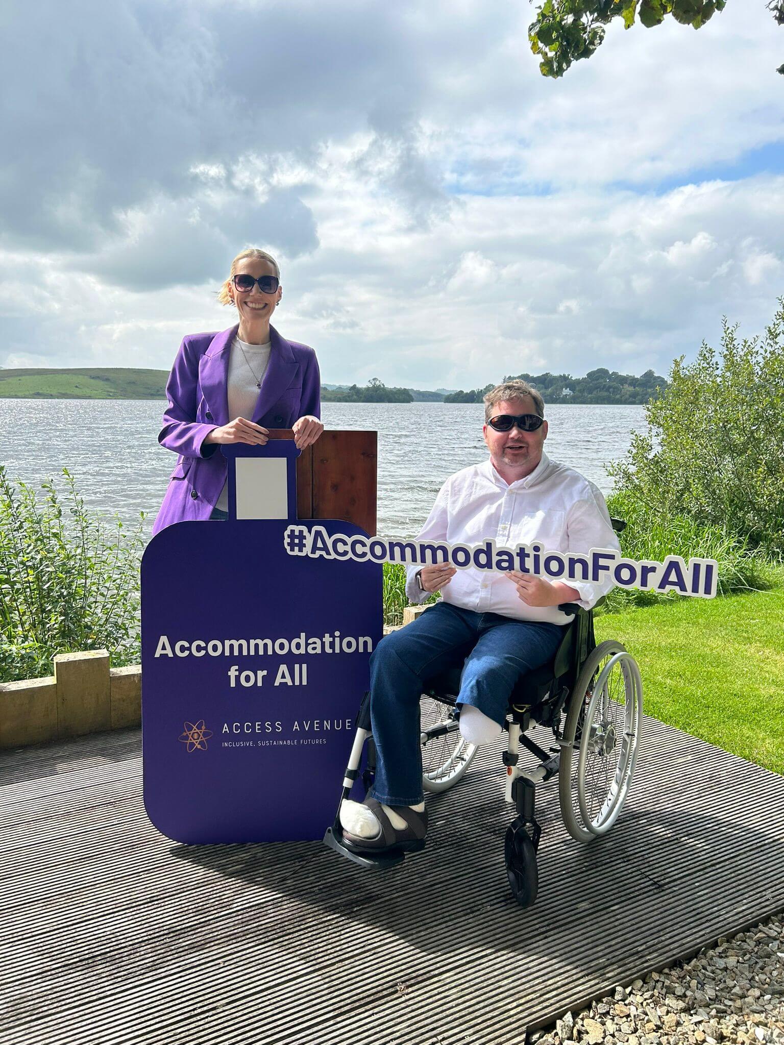 "Outdoors by a lake. A woman in a purple blazer stands behind a podium with 'Accommodation for All' written on it. Next to her, a man in a wheelchair holds a sign reading '#AccommodationForAll'. Both are smiling. They're on a wooden platform surrounded by grass and shrubs. The background shows a lake and cloudy sky. The podium has the 'Access Avenue' logo, indicating an event or initiative focused on accessibility."