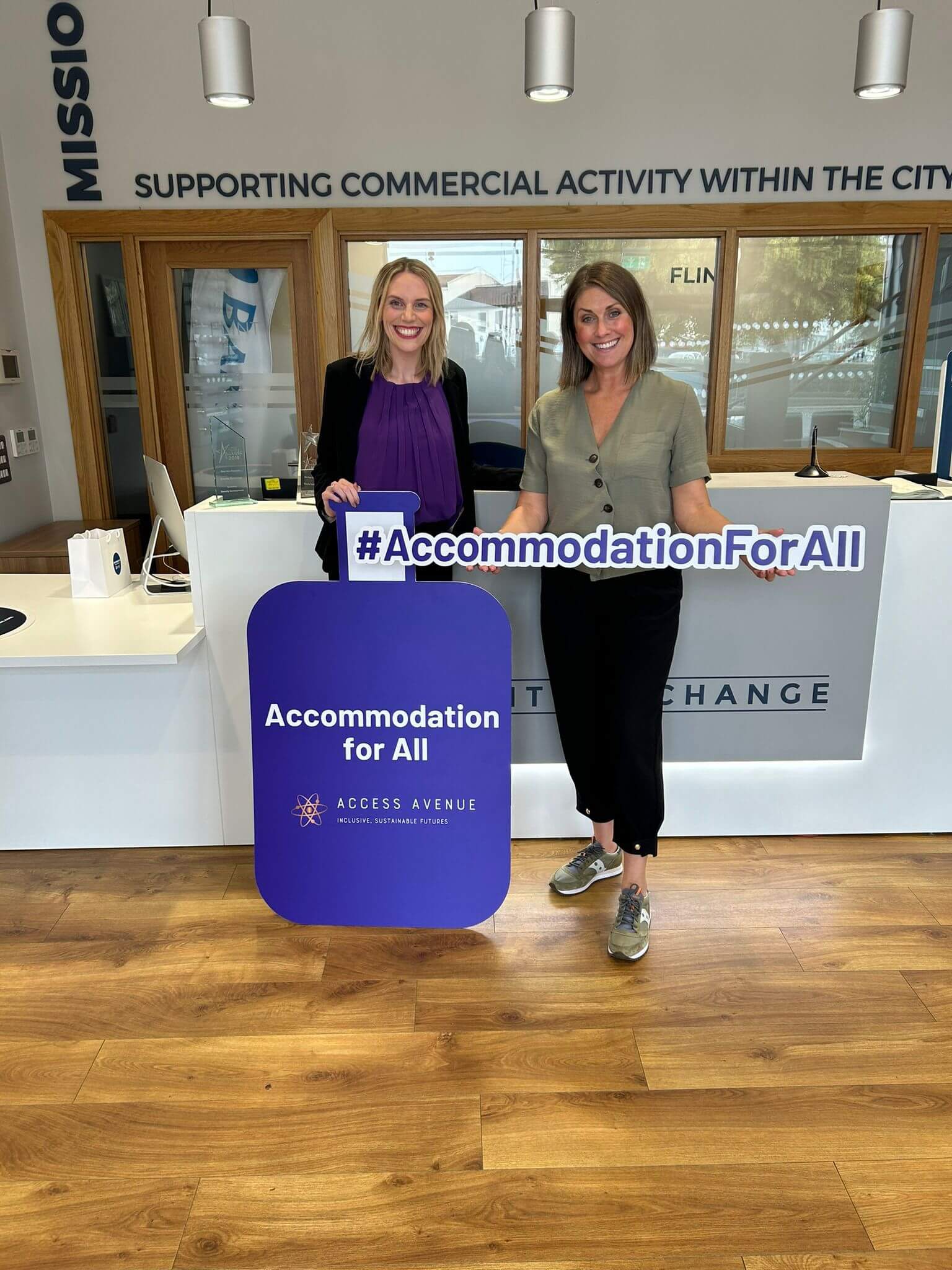 "Two smiling women stand in an office space. They're holding signs that read '#AccommodationForAll' and 'Accommodation for All' with the Access Avenue logo. The woman on the left wears a purple top, while the one on the right wears a green shirt. Behind them is a counter with 'EXCHANGE' visible. The wall above displays text: 'SUPPORTING COMMERCIAL ACTIVITY WITHIN THE CITY'. The room has wooden floors and spotlights on the ceiling."