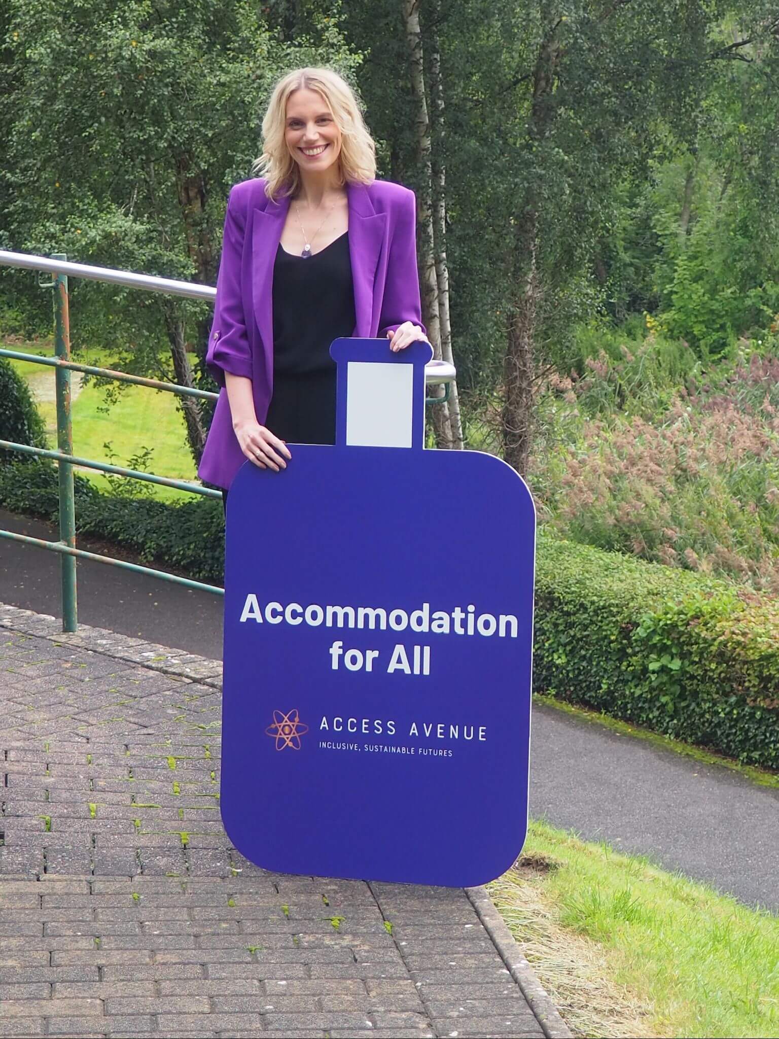 "A smiling blonde woman in a purple blazer stands on a pathway outdoors. She's holding a large purple sign that reads 'Accommodation for All' with 'Access Avenue' and a logo underneath. The background shows a wooded area with trees and greenery. There's a metal railing visible beside the path, suggesting it might be accessible."