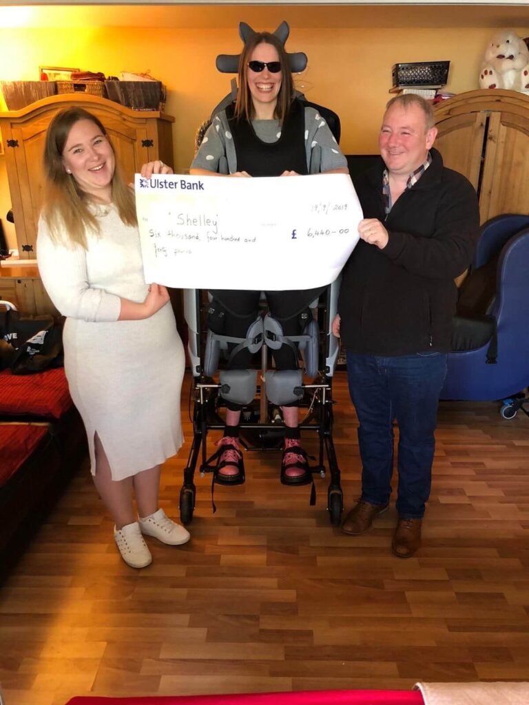 Three people are standing and smiling while holding a large check from Ulster Bank. The woman in the center is using a mobility standing frame and wearing sunglasses, with her legs secured in the frame. She is flanked by two people, one on each side— a woman in a white dress and a man in a black jacket. The check is made out to 'Shelley' for the amount of £6,460.00, and the setting is a cozy room with wooden flooring and light-colored furniture.