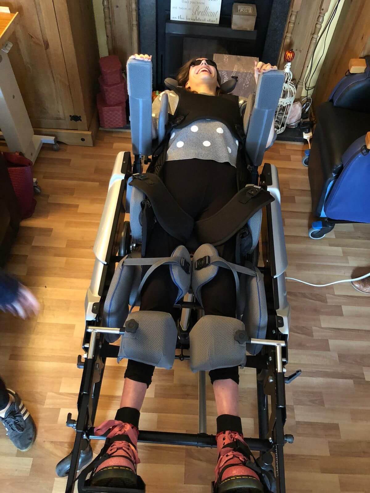 A woman is lying back in a supportive mobility standing frame with her arms raised in a joyful gesture. She is wearing sunglasses, a gray sweater with polka dots, and leg braces, with her feet secured in the frame. The room has a warm, cozy feel with wooden flooring, and various items are placed around, including some furniture and personal belongings.