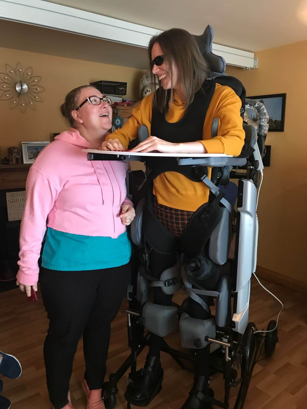 Two women are smiling and engaging in conversation. One woman is wearing a pink and teal hoodie, while the other is using a mobility standing frame with leg supports and wearing sunglasses. The atmosphere appears warm and supportive, with a cozy indoor setting, featuring wooden floors and soft lighting