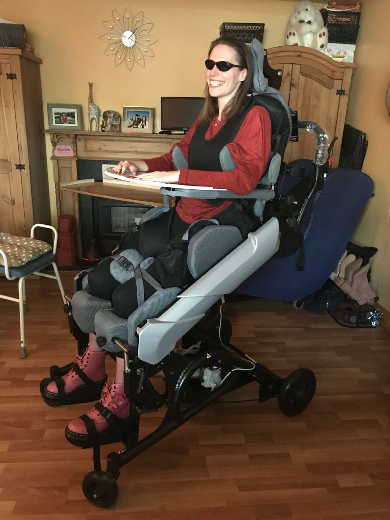 A person with a disability is reclining in a motorized wheelchair in their living room. The wheelchair has a headrest and footrests, allowing the person to lie down comfortably. There is a TV mounted on the wall above the wheelchair