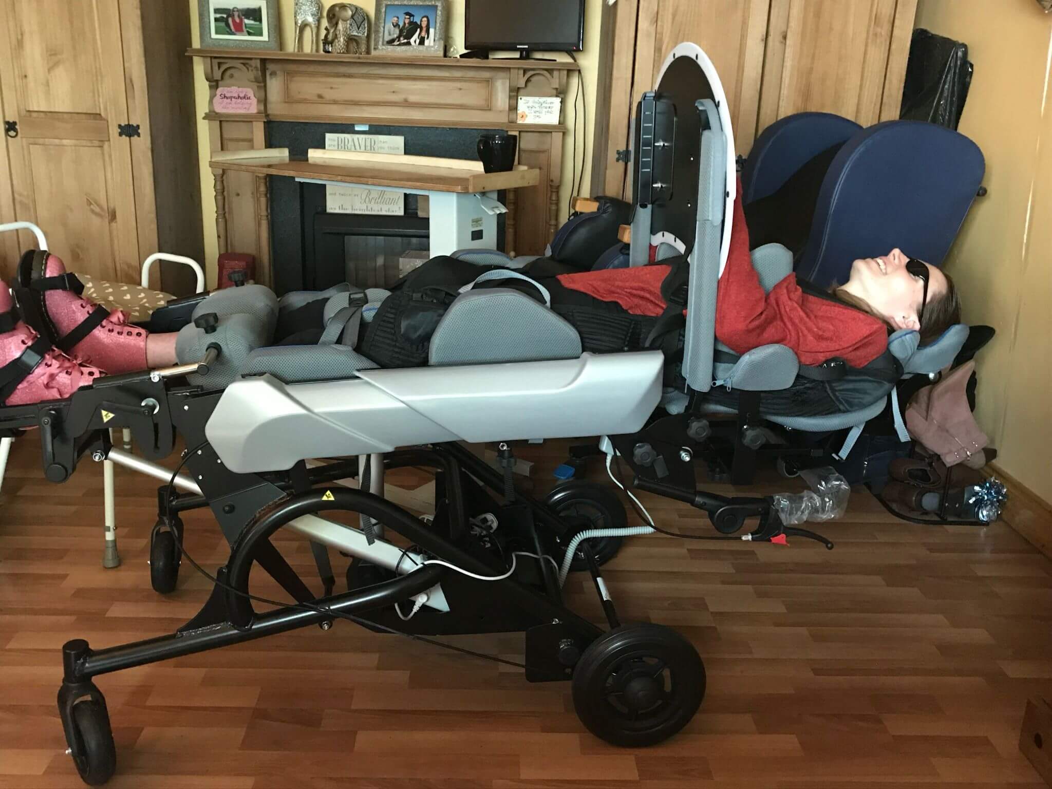 Shelley with a disability is reclining in a motorized wheelchair in their living room. The wheelchair has a headrest and footrests, allowing the person to lie down comfortably. There is a TV mounted on the wall above the wheelchair