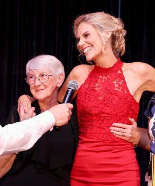 A group of four people are standing in front of a dark background. They are all smiling and looking at the camera. The person on the left is a man in a white shirt and black vest. He is holding a microphone. The person in the center is a woman with short gray hair and a black dress. She is holding a microphone. The person on the right is a woman with long blonde hair and a red dress. She is holding a microphone. The person on the right is a woman with long blonde hair and a blue and white patterned dress. She is holding an award. The award is a gold and blue plaque with a heart shape cut out of the center. The plaque states "Monthly Reporter Community Champion Awards 2024" and is presented by an organization called "encirc". The image is likely taken at an awards ceremony where the woman in the red dress is receiving an award for being a community champion.