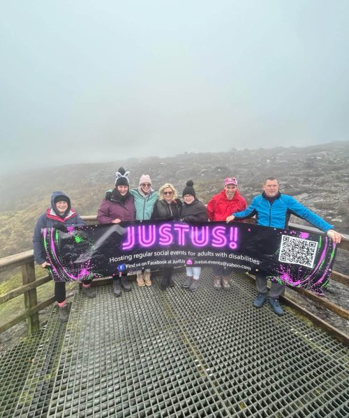 A group of seven people standing on a metal platform or walkway in a foggy, rocky landscape. They are holding a large purple and black banner that reads 'JUSTUS!' with the subtitle 'Hosting regular social events for adults with disabilities'. The group members are dressed in warm outdoor clothing of various colors.