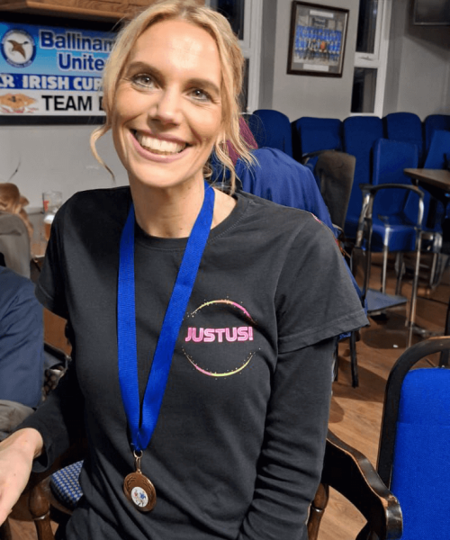 "A smiling blonde woman wearing a black shirt with a 'JUSTUS!' logo is seated in what appears to be a meeting room or club house. She's wearing a blue lanyard with a medal. In the background, there are blue chairs and a banner that reads 'Ballina United' and mentions an Irish team. The woman appears to be at a sports or community event."